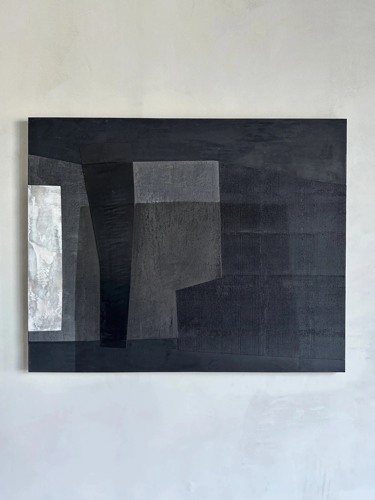 Charcoal geometric relief of woven and ribbed blocks surrounding one bright silver panel, on a cream wall.