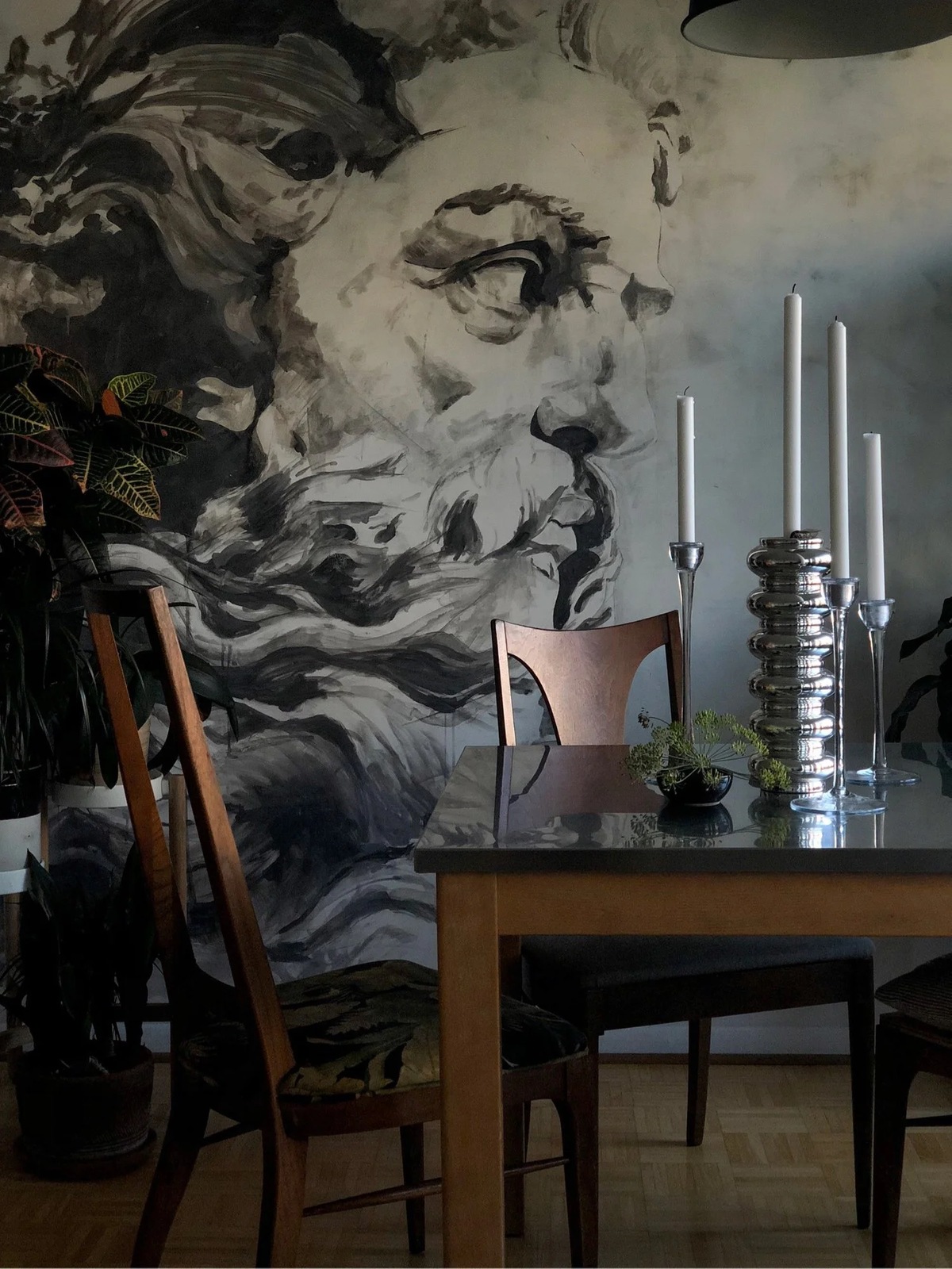 A monochrome figurative mural of a classical bearded head in charcoal and bone tones filling a dining room wall, silver candlesticks on the table below.