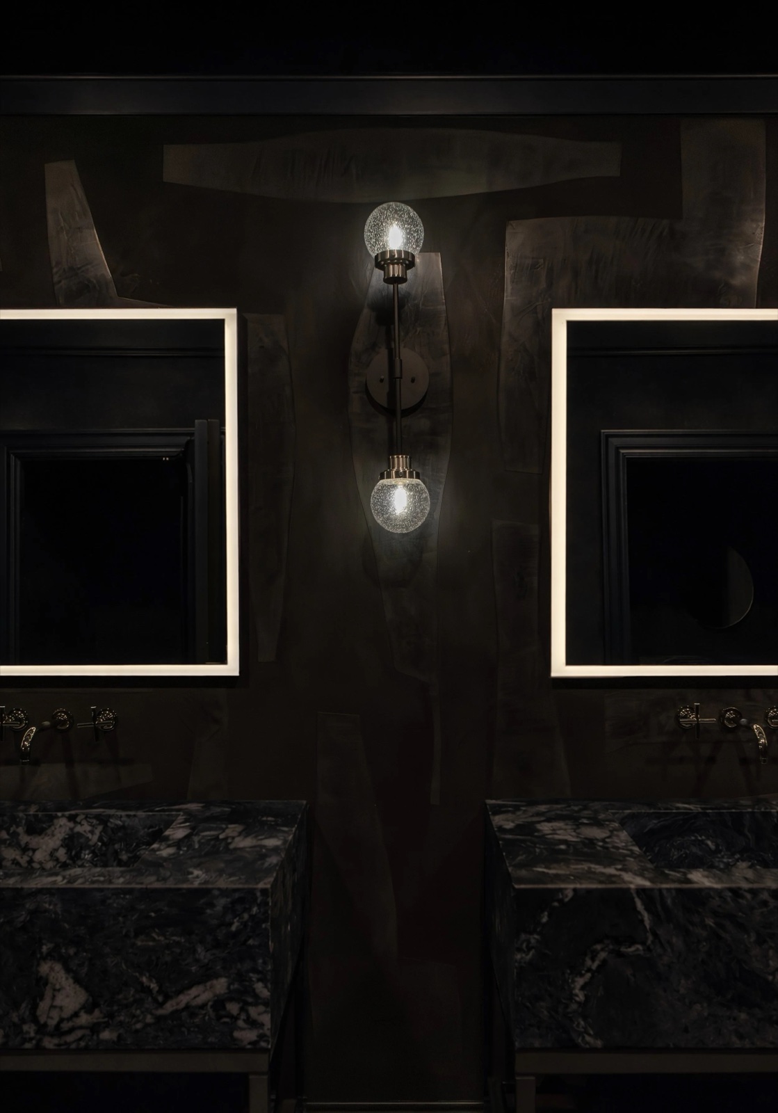 A dark powder room with a black sculptural plaster panel between two backlit mirrors and a brass sconce.
