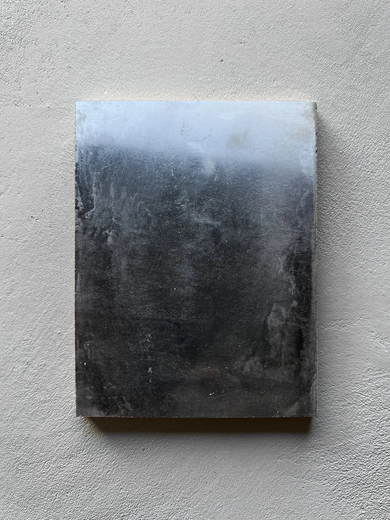 A small rectangular finish-sample panel of high-gloss Venetian plaster mounted on a light gray textured wall, showing a cloud-like tonal gradient from silver-blue to black.